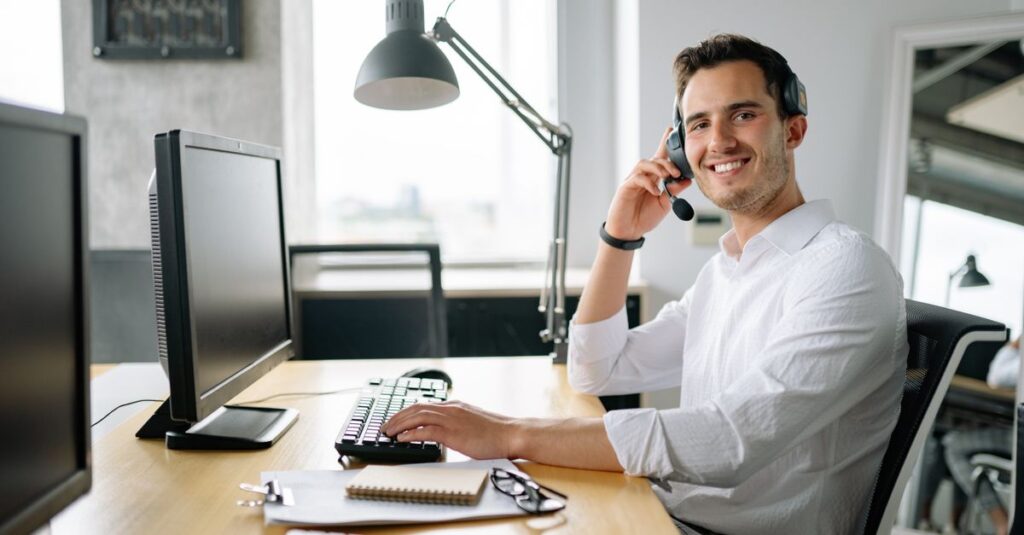 Smiling call center agent working at a desk with a headset and computer in a modern office.